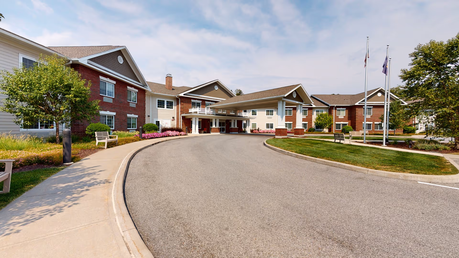 Front exterior view of The Landing of Poughkeepsie senior living facility showing a curved driveway, landscaped greenery, benches, and flagpoles under a partly cloudy sky.