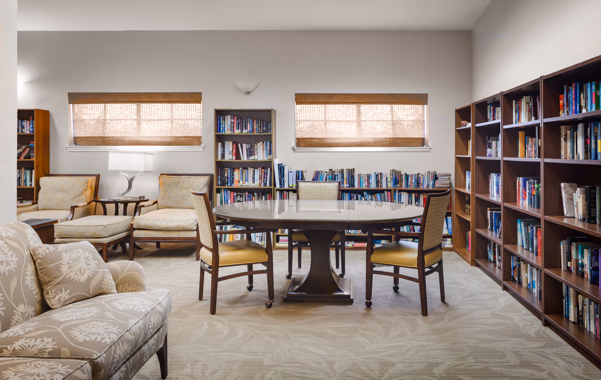 A cozy reading room with bookshelves filled with books along the walls, a round wooden table with four chairs in the center, two armchairs with matching ottomans near a side table with a lamp, and a patterned upholstered sofa in the foreground. The room has two windows with woven shades and light-colored walls and carpet.