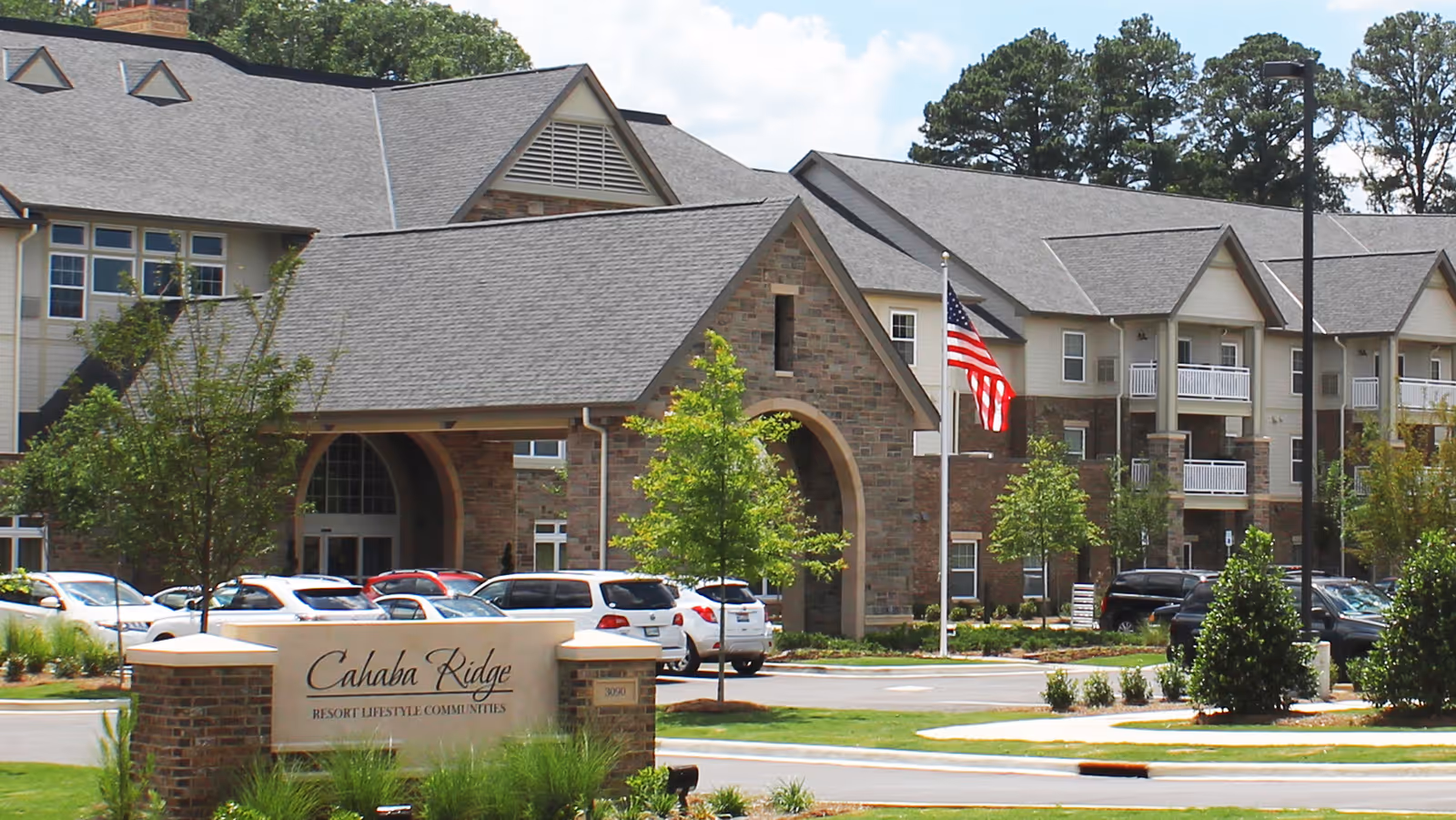 Exterior view of Cahaba Ridge Retirement Community building with multiple floors, a covered entrance, parked cars, trees, and an American flag on a flagpole.