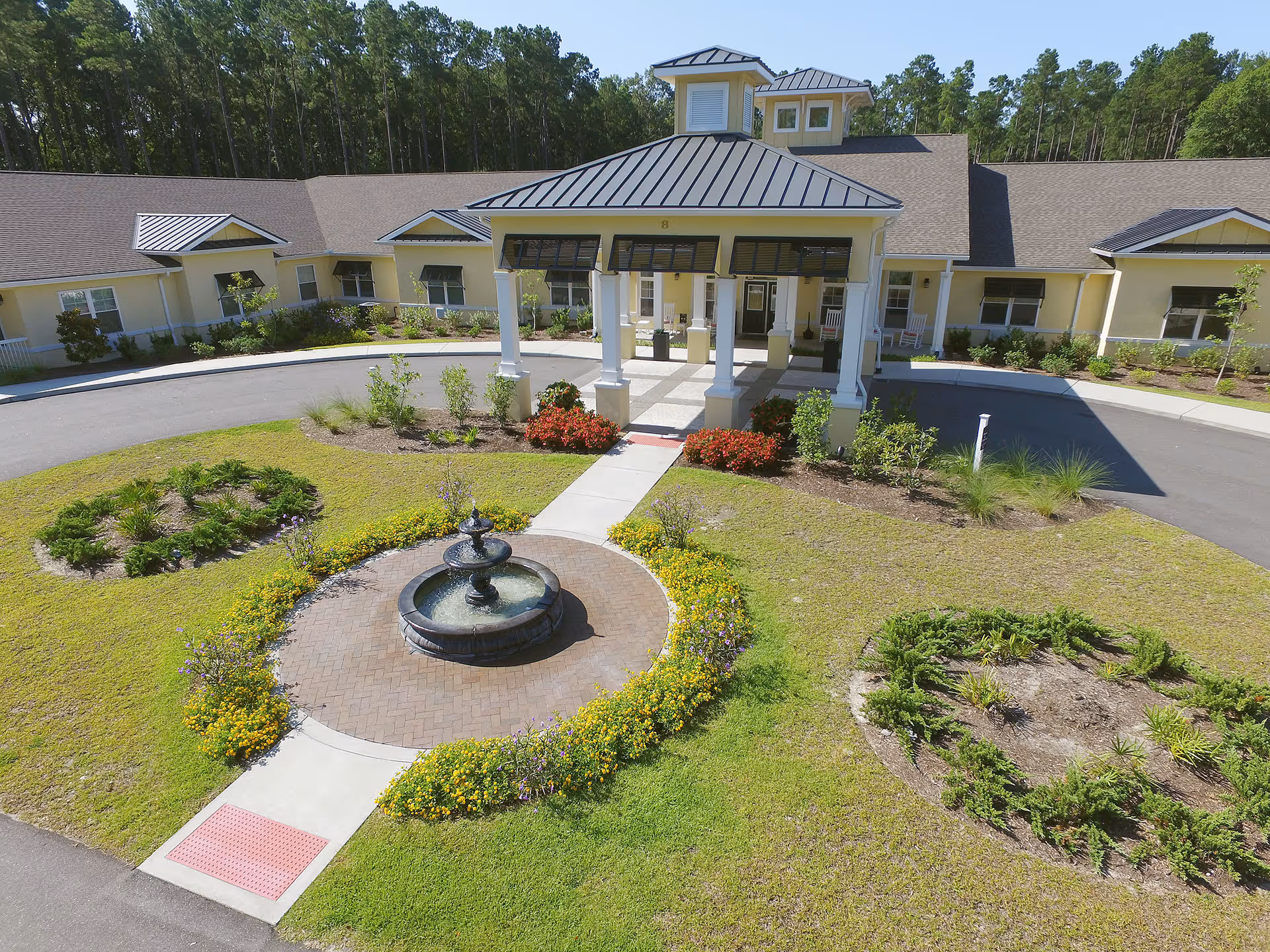 Front exterior view of a single-story senior living facility with a covered entrance, circular driveway, landscaped garden beds, and a central water fountain surrounded by flowers and greenery.