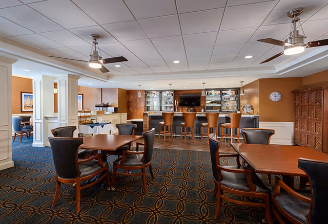 Interior view of a senior living facility dining area with wooden tables and chairs arranged on a patterned carpet. In the background, there is a bar with four high chairs, glass shelves with bottles and glasses, and a wall-mounted TV. The room has a white ceiling with recessed lighting and ceiling fans, and warm-toned walls with a clock and decorative elements.