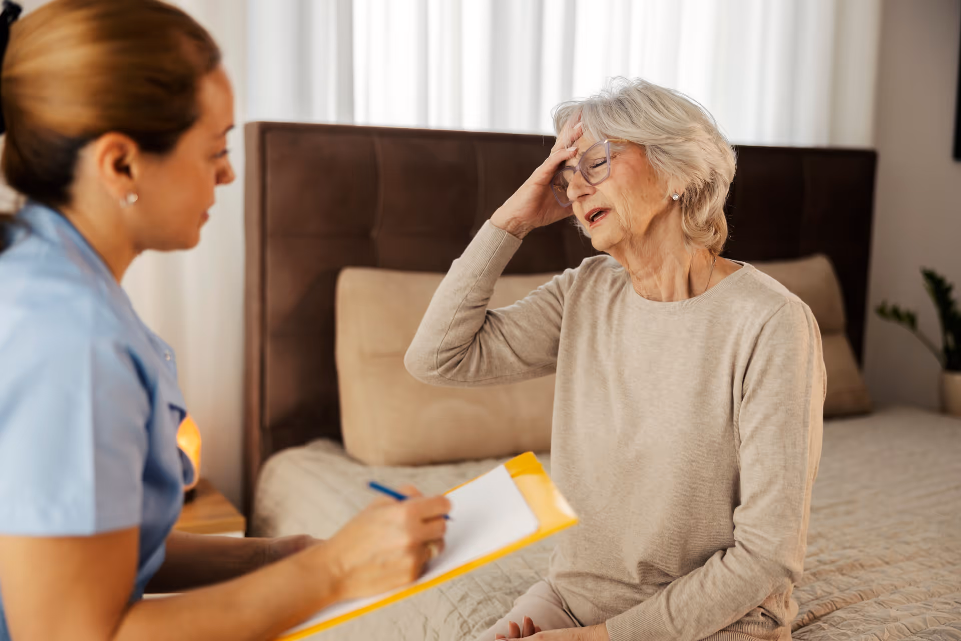 An elderly woman sitting on a bed with her hand on her forehead, appearing distressed or unwell, while a healthcare professional in blue scrubs takes notes on a clipboard beside her.