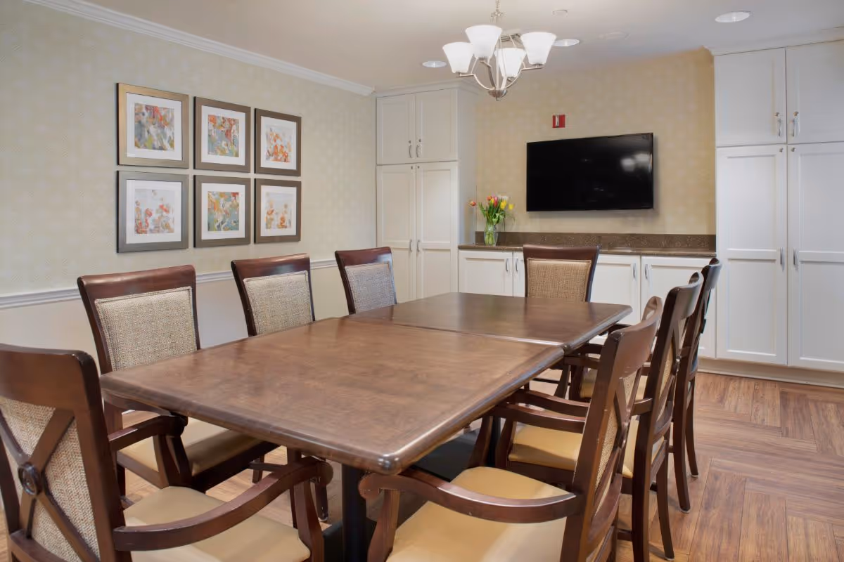 A dining room with a large wooden table surrounded by eight wooden chairs with cushioned seats and backs. The room has light-colored wallpaper, a chandelier hanging from the ceiling, a flat-screen TV mounted on the wall, white cabinets, and framed artwork on the wall.