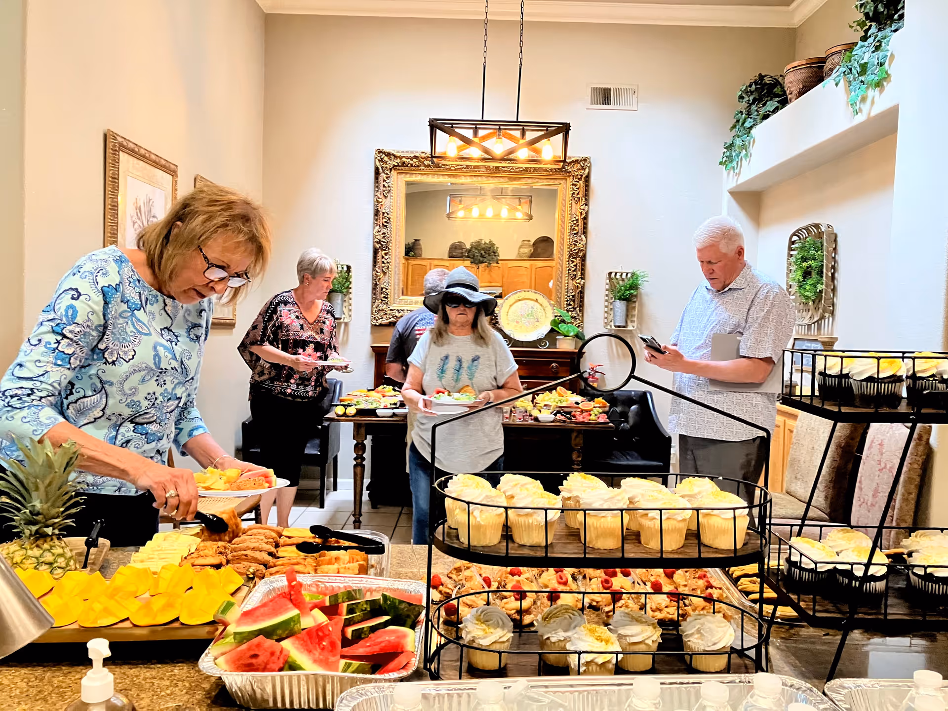 Several elderly people serving themselves food from a buffet table inside a well-lit room with decorative plants and a large ornate mirror on the wall.