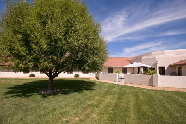 A large green tree stands on a well-maintained grassy lawn in front of a single-story building with a red-tiled roof and light-colored walls. The building has a small patio area with a white umbrella and some plants. The sky is blue with some wispy clouds.