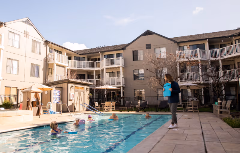 Outdoor swimming pool area at Baywood Court with several elderly people in the pool using kickboards and a staff member standing poolside holding a kickboard. The pool is surrounded by a multi-story residential building with balconies, patio furniture, umbrellas, and some leafless trees under a partly cloudy sky.