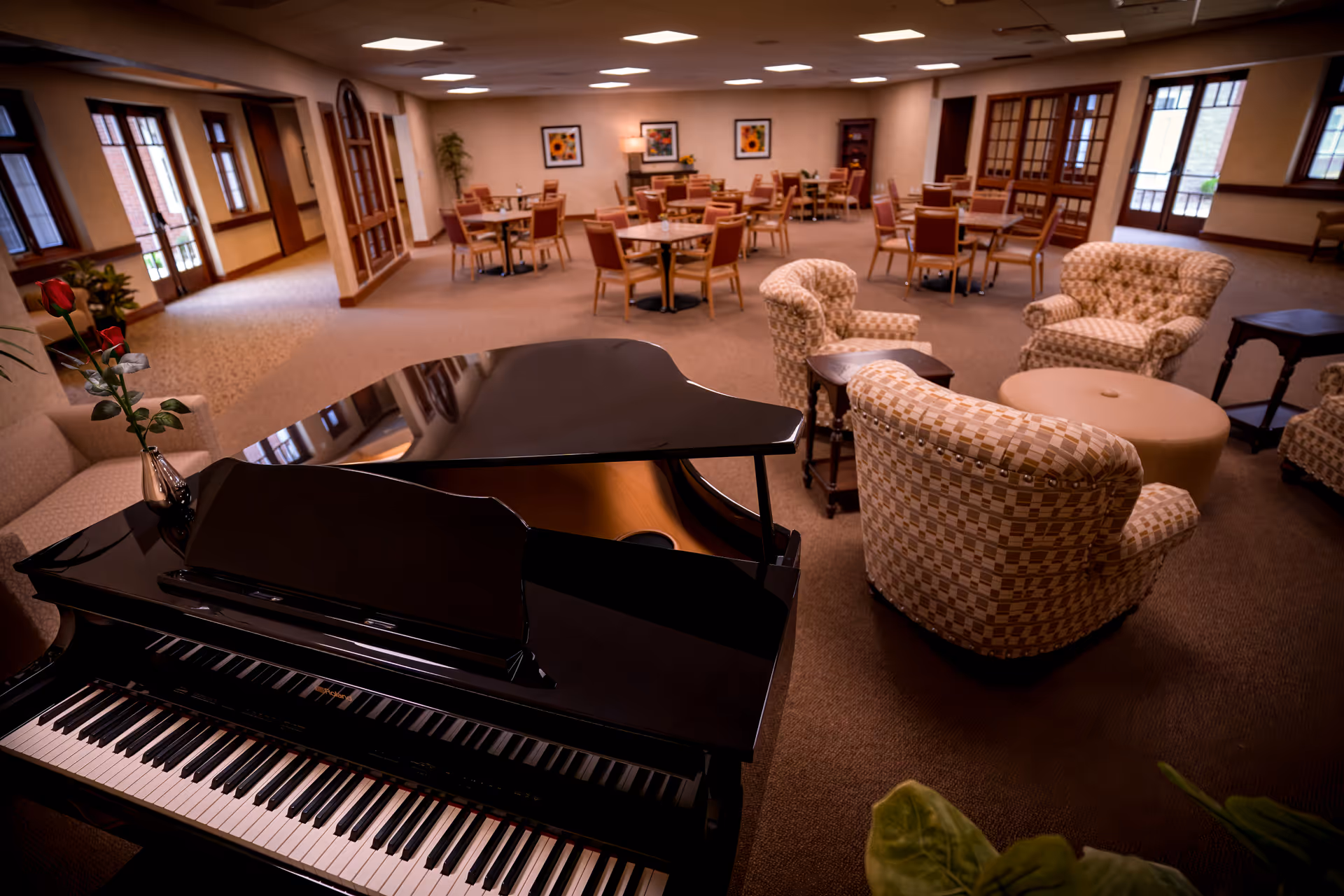 A spacious and well-lit common area featuring a black grand piano with a single red rose in a vase on top, several upholstered armchairs arranged around a round ottoman, and multiple dining tables with chairs in the background. The room has large windows and glass doors allowing natural light to enter, with framed artwork on the walls and carpeted flooring.