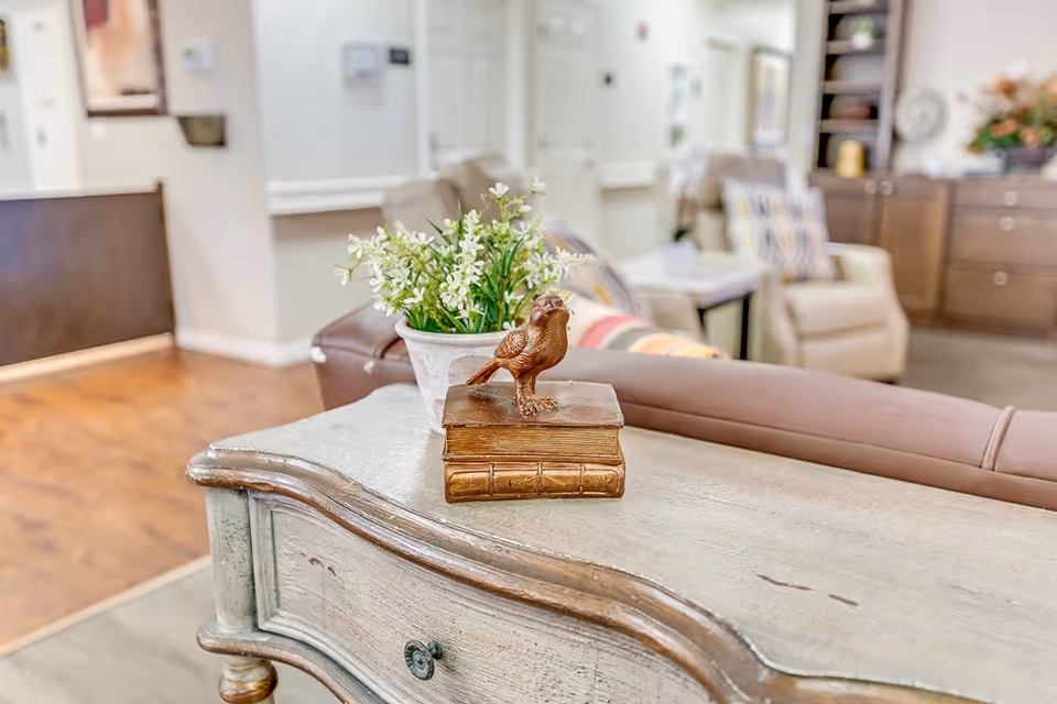 A cozy senior living common area with a wooden table in the foreground holding a decorative bird statue on top of two books and a potted plant with white flowers. In the background, there are comfortable armchairs with patterned cushions, a side table, and cabinetry with decor items.