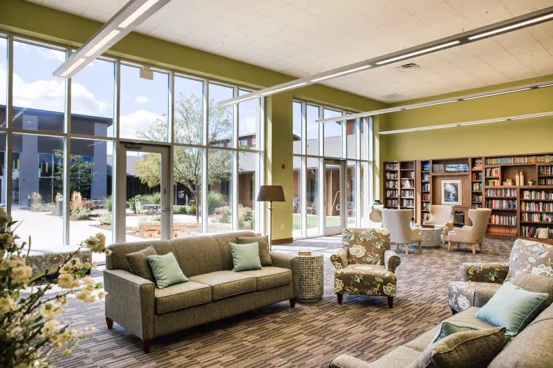 A bright and spacious senior living community lounge with large floor-to-ceiling windows letting in natural light. The room features comfortable sofas and armchairs with cushions, a floor lamp, and a bookshelf filled with books. Outside the windows, a landscaped courtyard with trees and plants is visible.
