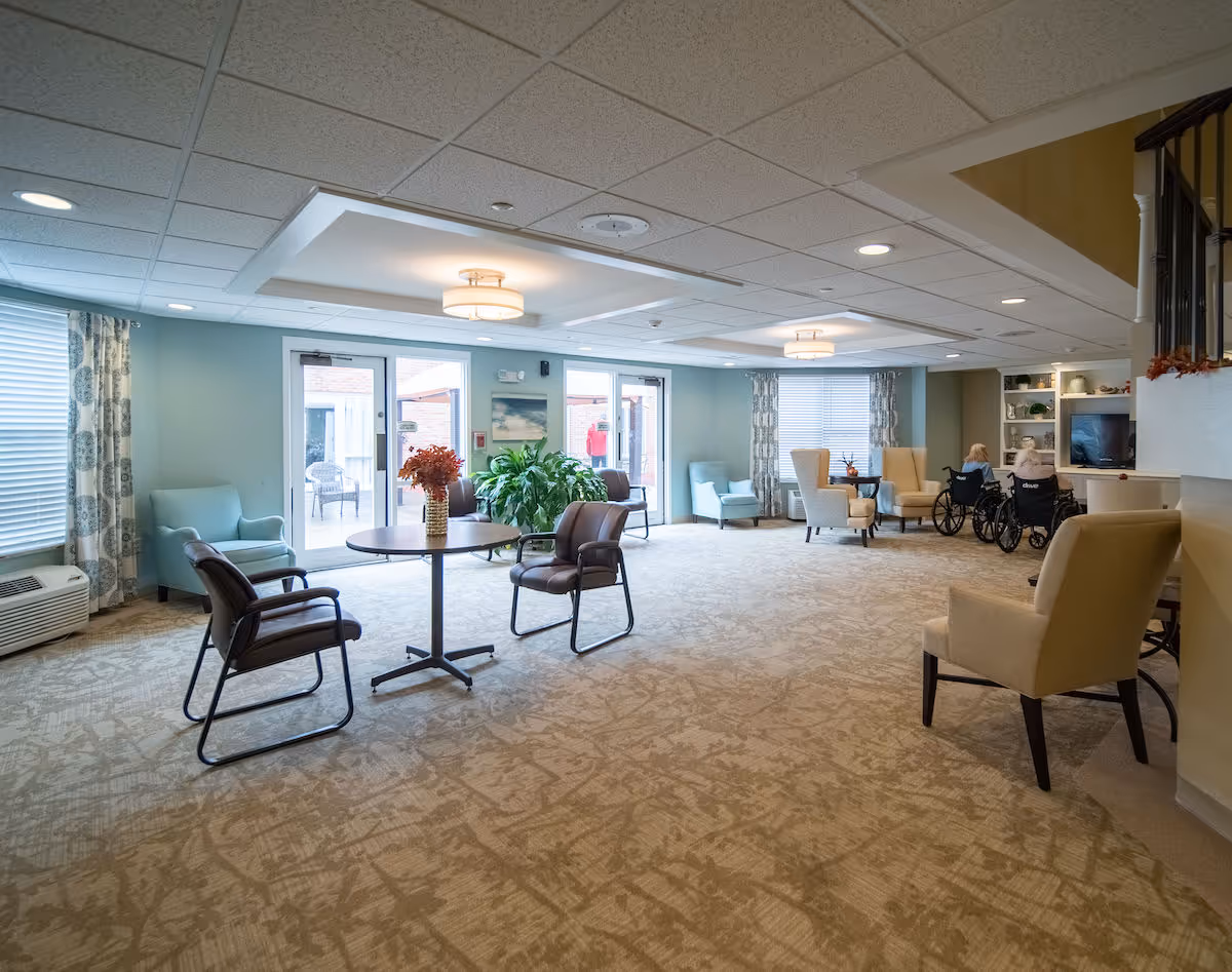 Spacious, well-lit senior living lounge with chairs, tables, plants, large windows and two people in wheelchairs near a TV.
