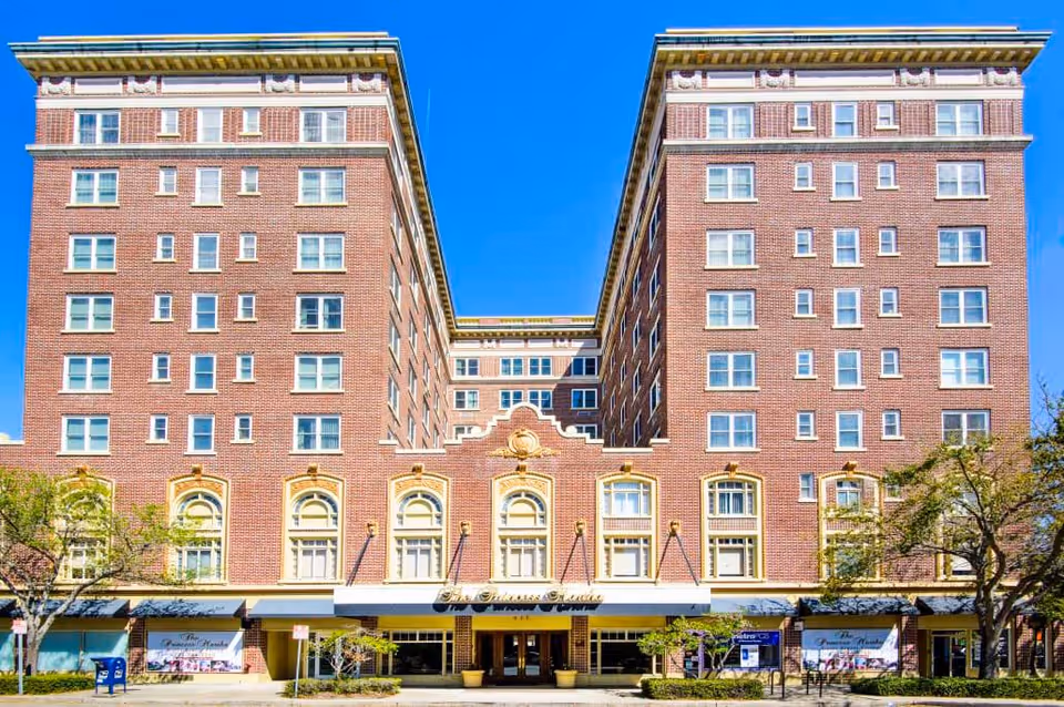 A brick senior living building with two tall wings and a central entrance against a bright blue sky.