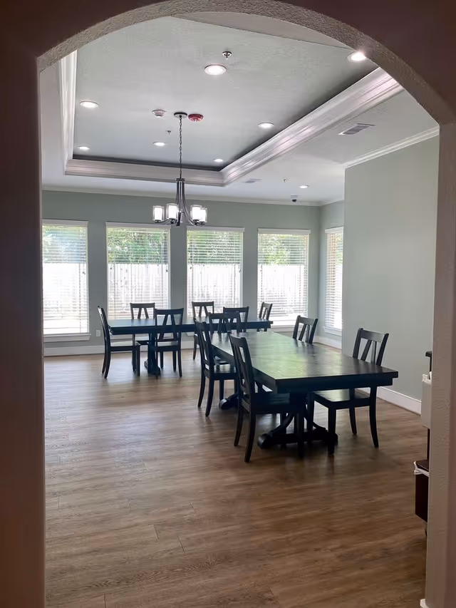 A dining room with two dark wooden tables and several matching chairs arranged around them. The room has large windows with white blinds letting in natural light, light green walls, and a ceiling with recessed lighting and a central chandelier. The floor is wood with a light brown finish.