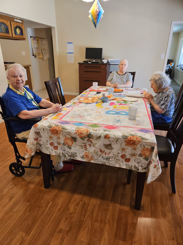 Three elderly women sitting around a rectangular dining table covered with a floral and colorful tablecloth. One woman is in a wheelchair on the left side, smiling at the camera. The other two women are seated on the opposite side and right side of the table, one looking forward and the other reading a book or notebook. The room has wooden flooring, beige walls, and a wooden cabinet with a computer monitor on top in the background.
