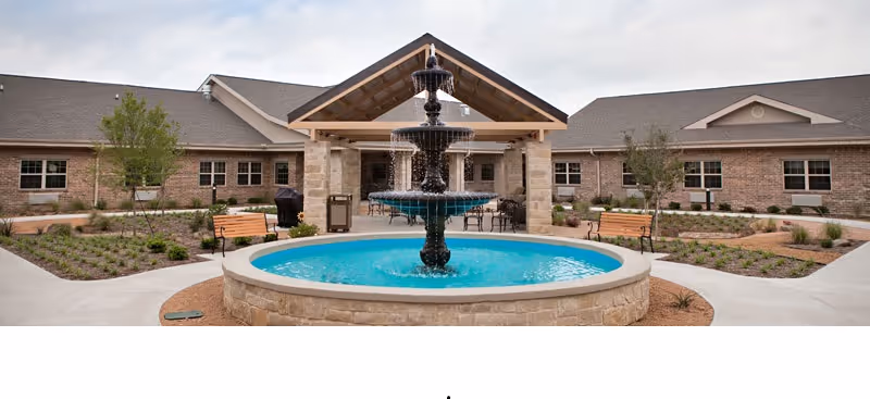 A circular fountain and benches in a landscaped courtyard in front of a single-story brick senior living facility with a covered entrance.