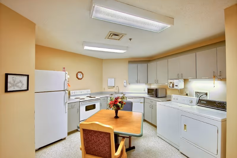 A well-lit kitchen area with white appliances including a refrigerator, stove, microwave, dishwasher, and a washing machine and dryer. There are beige walls, white cabinets, and a wooden table with a floral centerpiece and a chair in the center of the room.