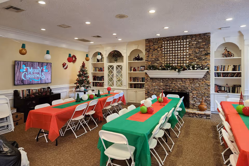 A festive decorated common room with tables covered in red and green tablecloths, white folding chairs, a stone fireplace adorned with garland, built-in bookshelves, a Christmas tree, and a TV screen displaying 'Merry Christmas'.