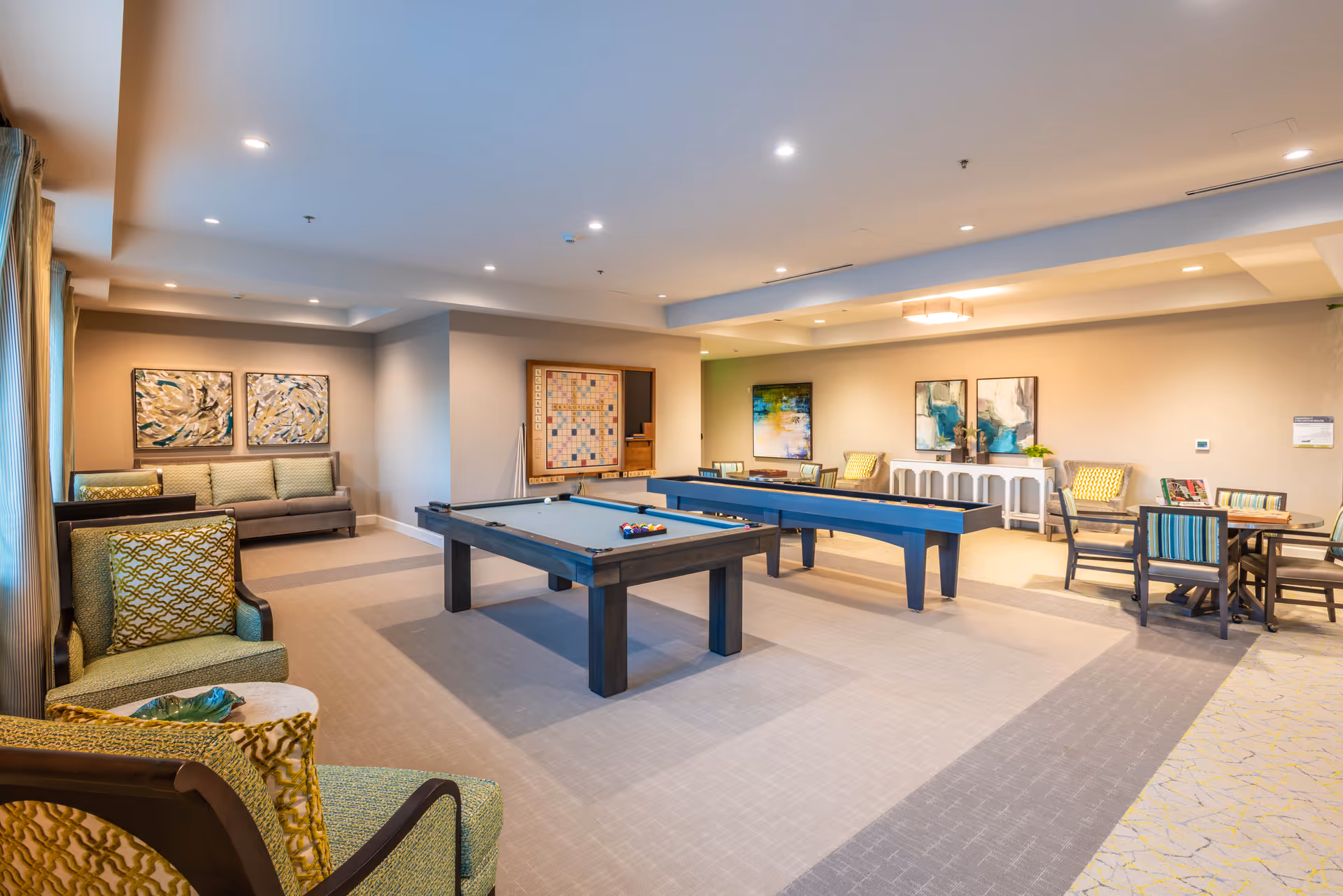 A spacious recreational room with a pool table and a shuffleboard table in the center. The room features comfortable seating including armchairs and sofas, several tables with chairs, and decorative artwork on the walls. The lighting is bright with recessed ceiling lights, and the room has a warm, inviting atmosphere.
