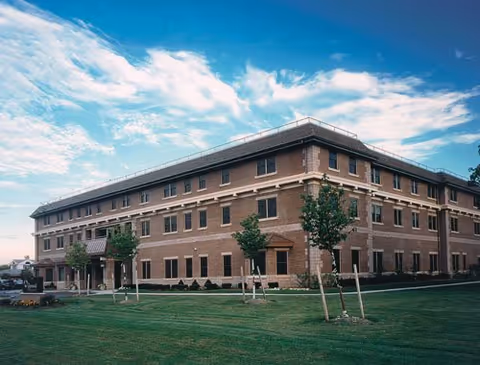 Three-story brick senior living facility building with a manicured lawn and small trees under a blue sky.