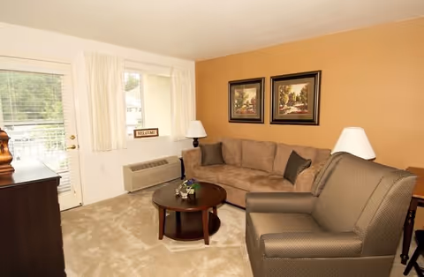 A cozy living room with a beige sectional sofa, a brown armchair, a round wooden coffee table with a small plant, two framed landscape paintings on a tan accent wall, a window with white curtains, a door with glass panels, and a small air conditioning unit below the window.