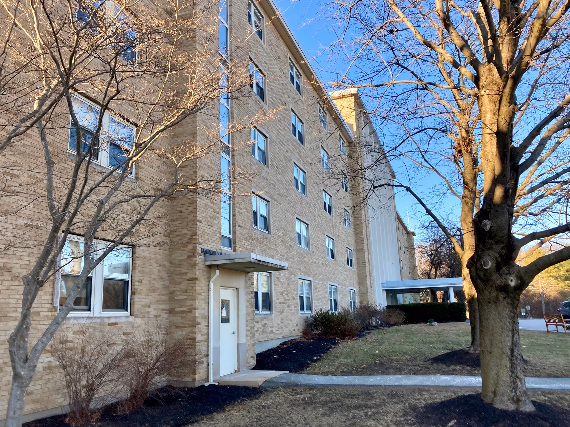 Exterior view of a multi-story brick building with several windows, a white door with a small awning, and leafless trees in the foreground. The sky is clear and blue, and there is a grassy area with mulch beds and a sidewalk leading to the entrance.