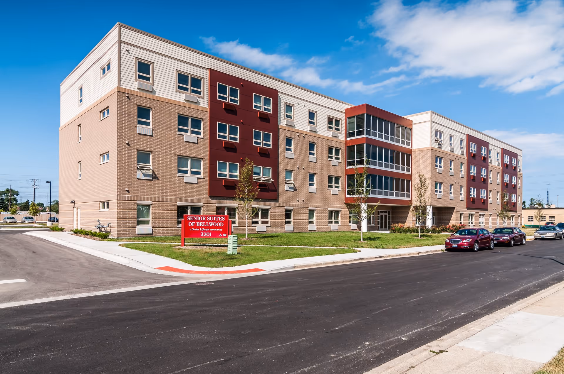 Exterior view of a modern four-story senior living facility named Senior Suites of Bellwood. The building features a combination of beige brick and white siding with red accent panels around some windows. There is a red sign in front of the building with the facility name and address, 3201. Several cars are parked along the street adjacent to the building, and the sky is partly cloudy.