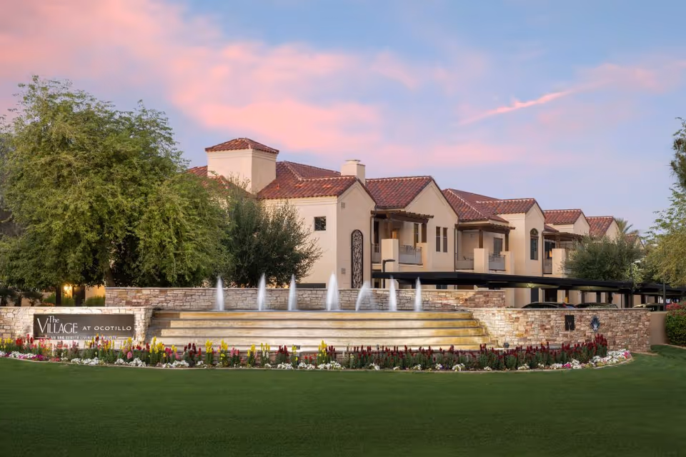 Exterior view of The Village at Ocotillo building with a tiered fountain, landscaped lawn, and flower beds at sunset.