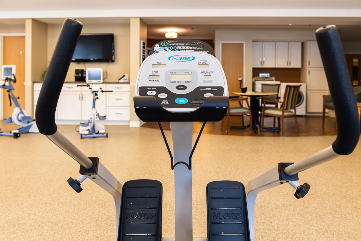 View of a NuStep exercise machine in a fitness or rehabilitation room with additional exercise bikes, a television mounted on the wall, and a seating area with a table and chairs in the background.