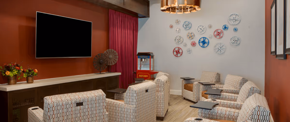 A cozy lounge area with patterned armchairs arranged in rows facing a large flat-screen TV mounted on a rust-colored wall. There is a popcorn machine in the corner and decorative wall art featuring colorful circular gears. The room has a warm and inviting atmosphere with a mix of modern and vintage decor elements.