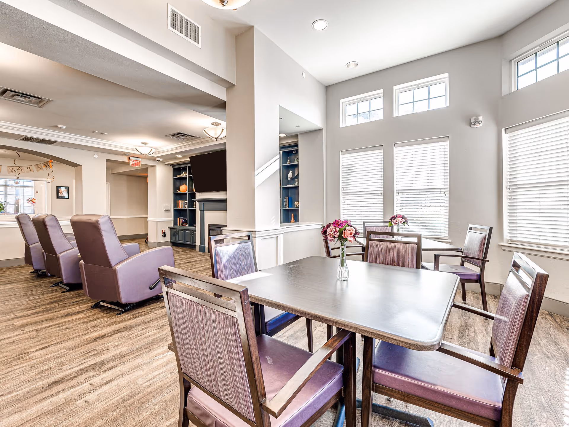 A bright and spacious common area in a senior living facility featuring a table with four chairs and a small vase of flowers on top. In the background, there are four purple reclining chairs facing a wall-mounted TV above a fireplace, with built-in shelves on either side. Large windows with white blinds allow natural light to fill the room, and the flooring is wood-style laminate.