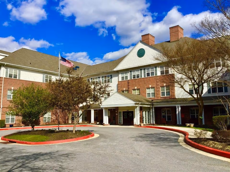 Two-story brick senior living building with entrance, drive, trees, and American flag.