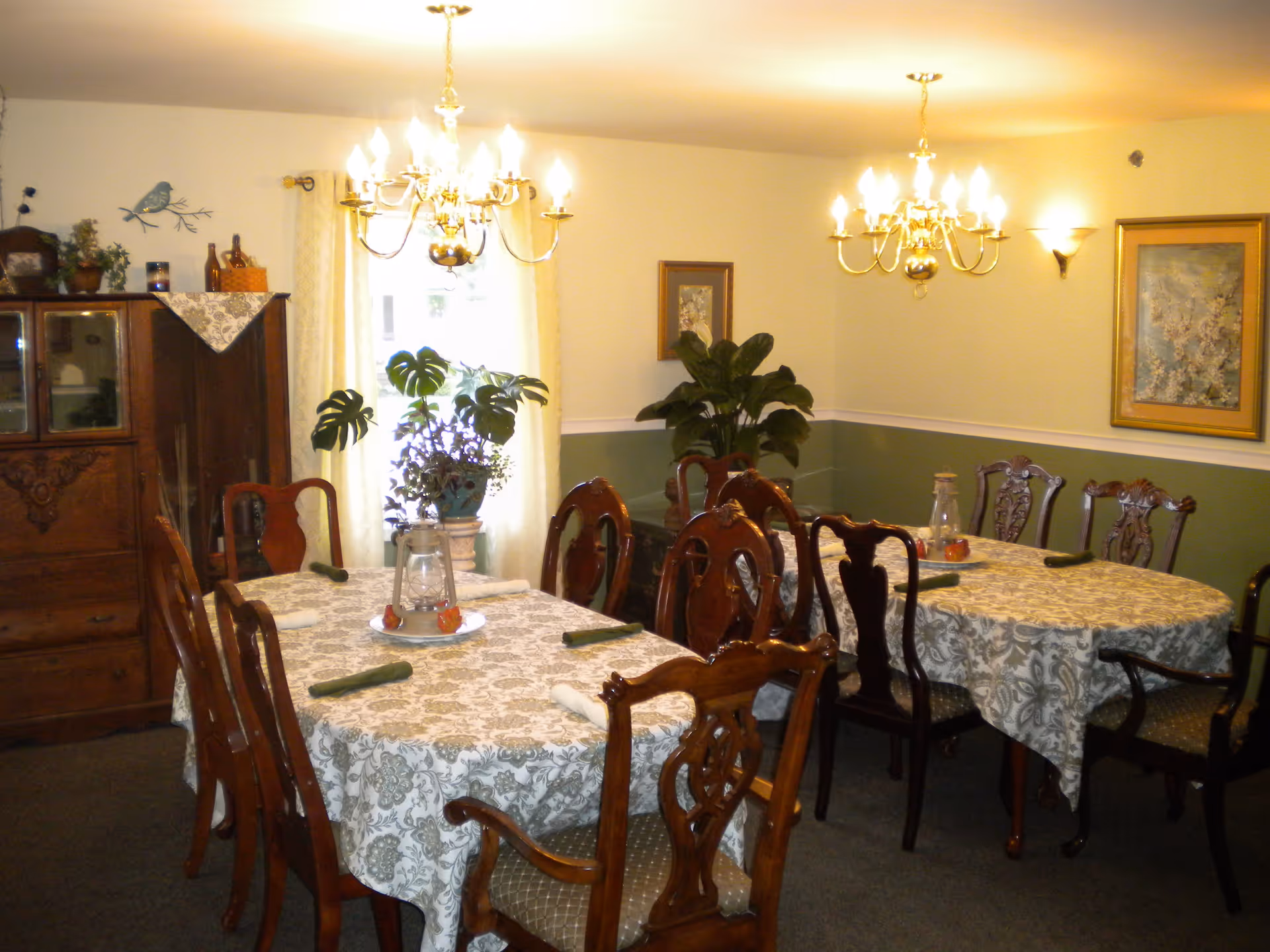 A dining room with two tables covered in patterned tablecloths, each surrounded by wooden chairs with cushioned seats. The tables have rolled green napkins and decorative centerpieces. The room is warmly lit by two chandeliers and has green and white walls adorned with framed artwork and plants. A wooden cabinet with glass doors is visible on the left side.