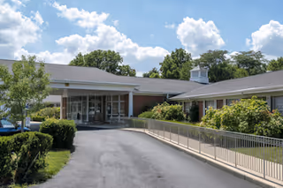 Exterior view of a single-story brick building with a covered entrance, surrounded by greenery and a paved driveway leading up to the entrance under a partly cloudy sky.