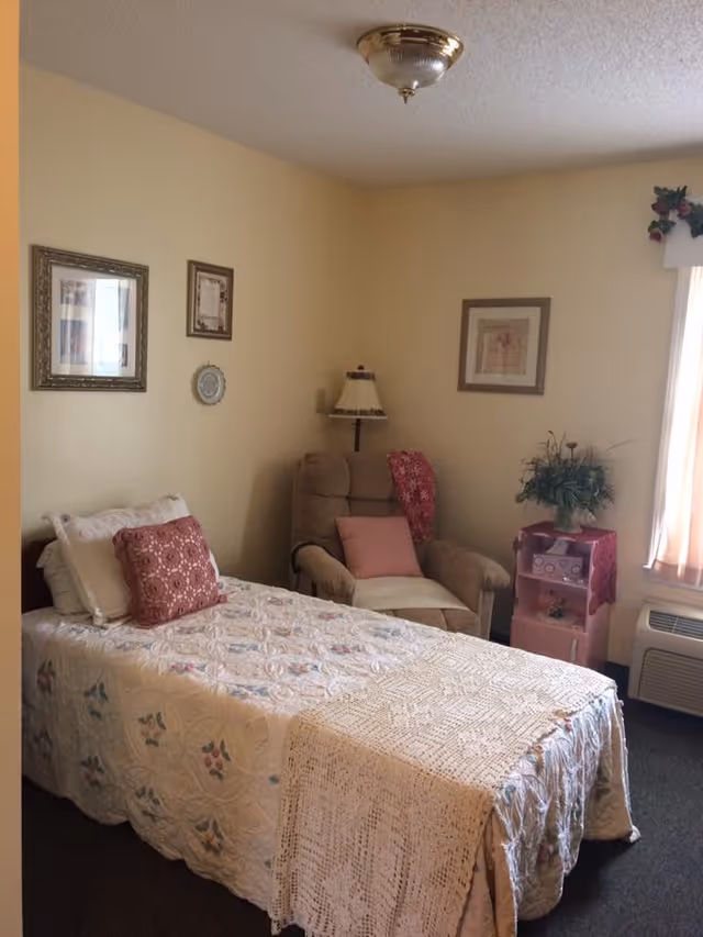 Small bedroom with a single bed covered by a quilt, an upholstered armchair, side table with a plant, and framed pictures on pale yellow walls.