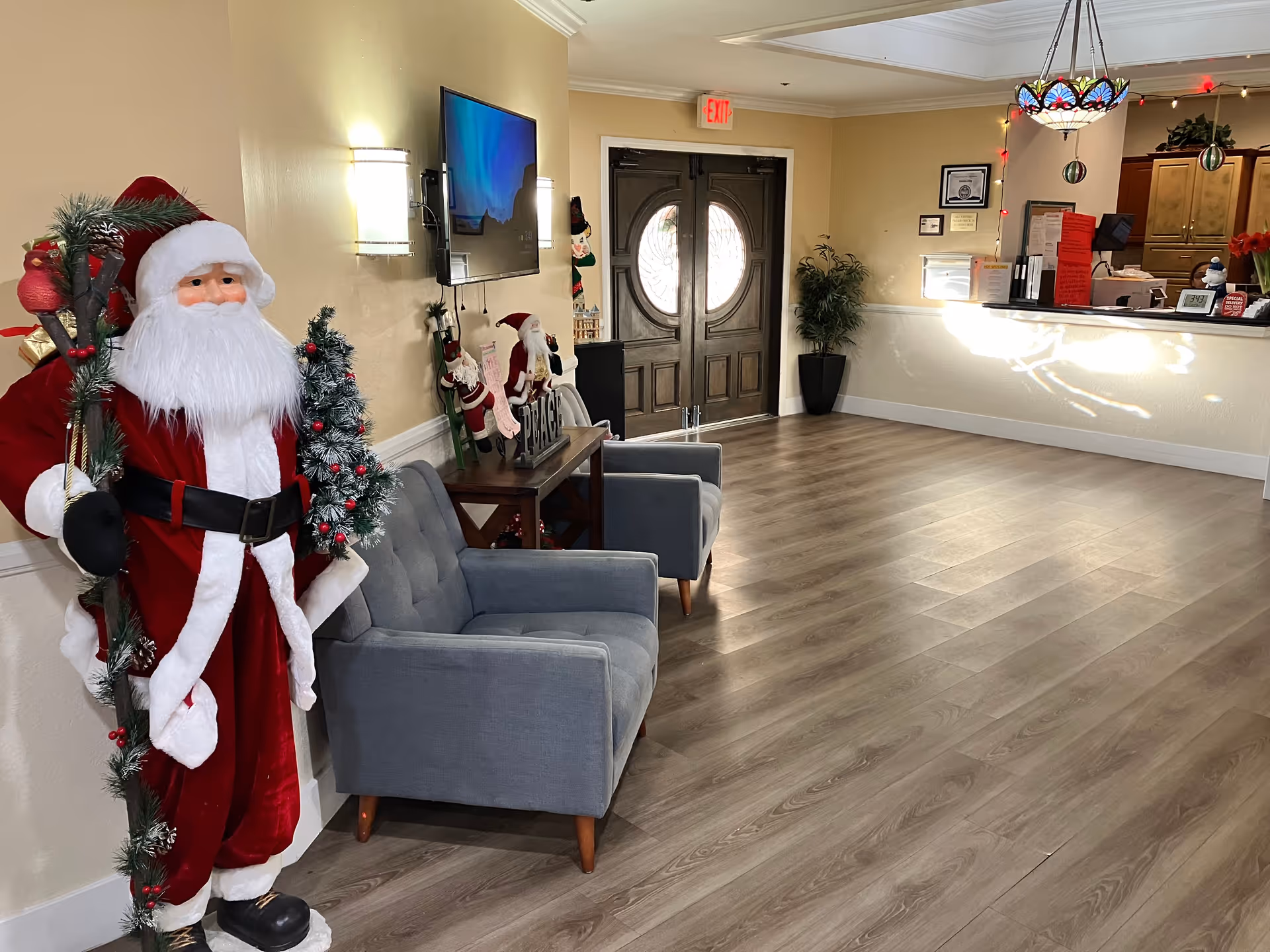 Interior view of a senior living facility lobby decorated for Christmas with a large Santa Claus figure holding a decorated staff, two gray armchairs, a wall-mounted TV, a small table with holiday decorations, double wooden doors with circular windows, a potted plant, and a reception desk with holiday ornaments and paperwork.