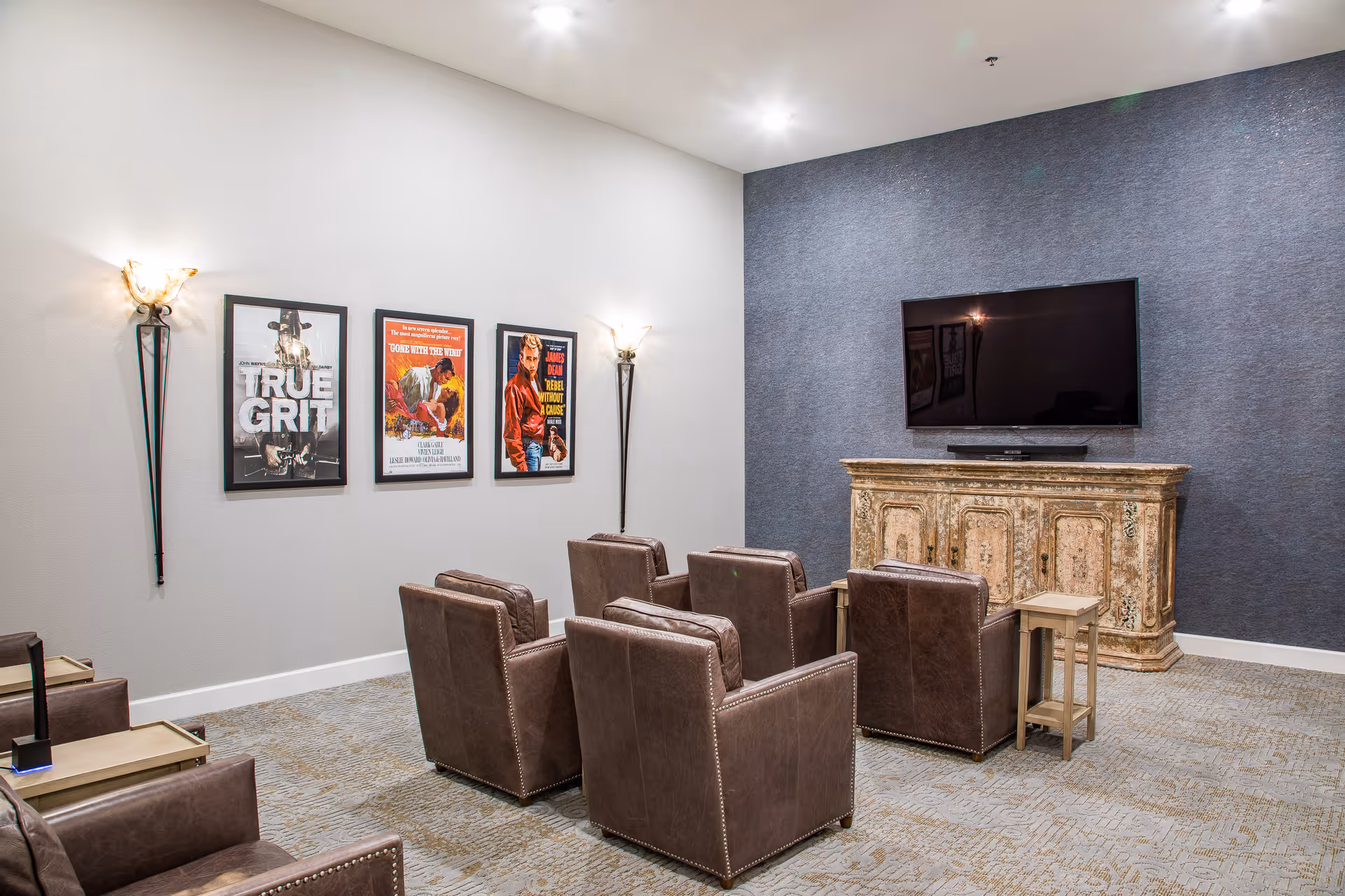 A cozy media room with six brown leather armchairs arranged in two rows facing a large flat-screen TV mounted on a textured blue-gray wall. Below the TV is an ornate wooden cabinet. The opposite wall is white with three framed movie posters and two wall-mounted sconces providing soft lighting. The floor is carpeted in a patterned design.