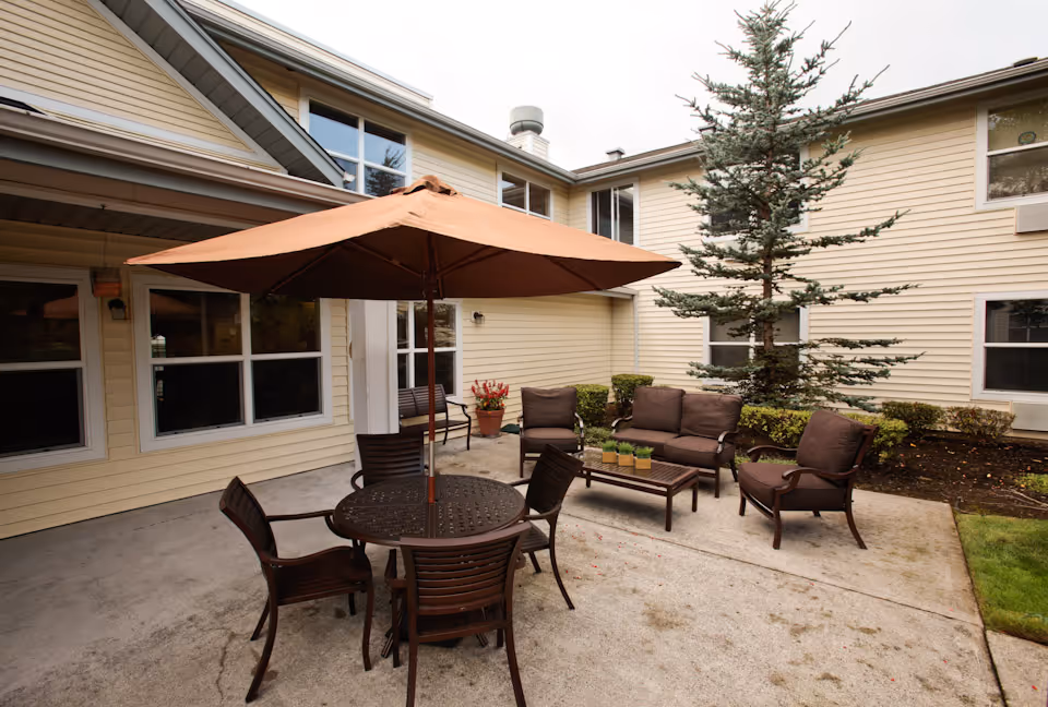 Patio courtyard with a round table under a large umbrella, several chairs and lounge seating in front of a light-yellow two-story building.