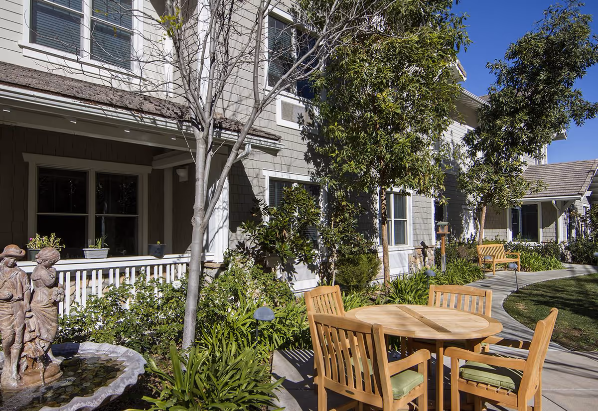Outdoor seating area at Ivy Park at Wood Ranch featuring a round wooden table with four chairs with green cushions, surrounded by trees, plants, and a curved concrete pathway. A small water fountain with statues is visible on the left side near the building with gray siding and white trim.