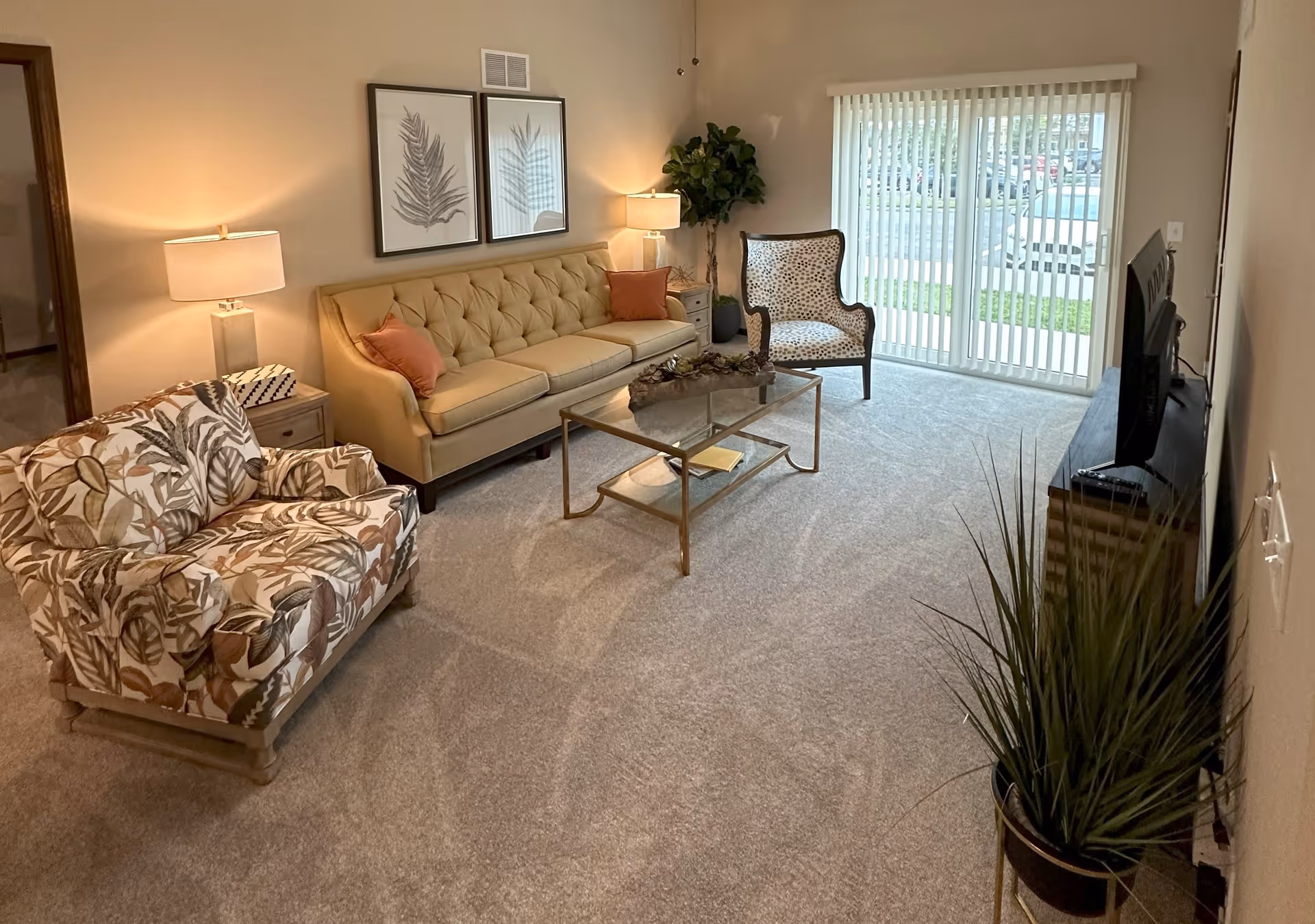 A cozy living room with a beige tufted sofa adorned with two orange pillows, a patterned armchair, and another armchair with a leaf design. A glass coffee table with a decorative centerpiece sits in the middle on a light carpeted floor. Two framed leaf prints hang on the wall above the sofa. There are two table lamps on side tables flanking the sofa, a potted plant in the corner, and a TV on a stand opposite the sofa. Large vertical blinds cover a sliding glass door that leads outside, where cars are visible in the parking lot.