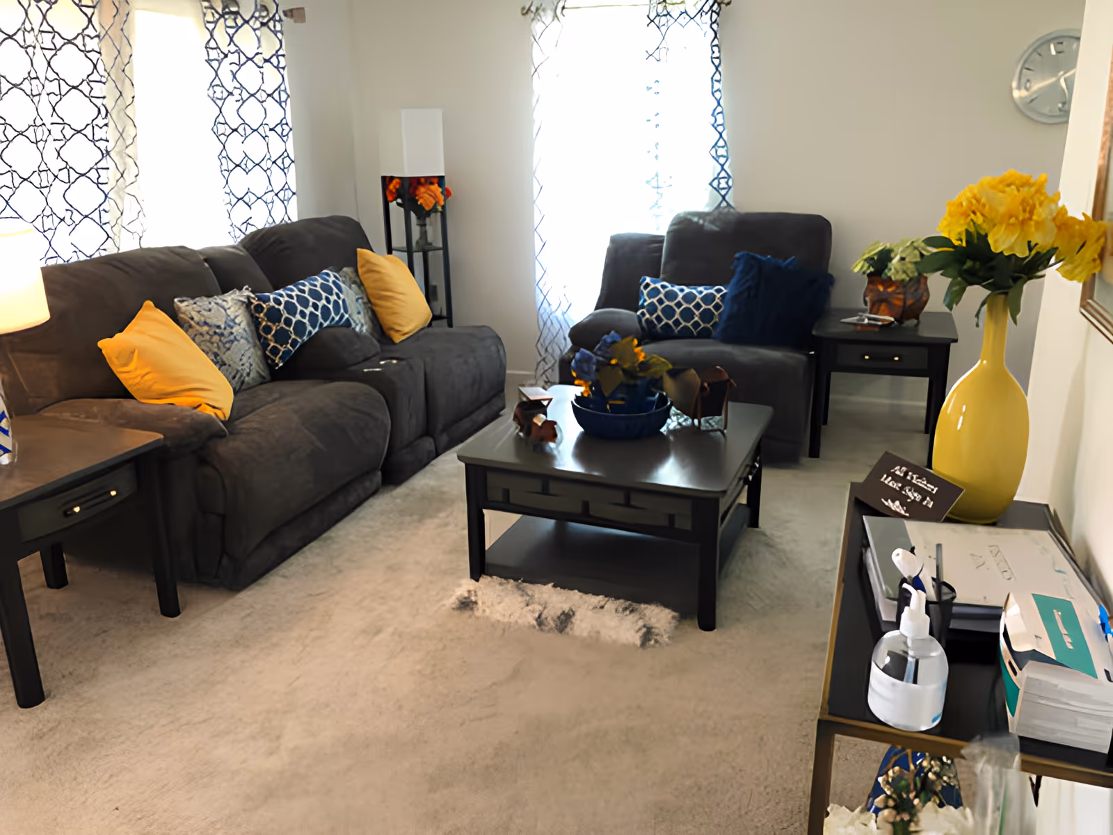 Cozy living room with dark gray sofas, blue and yellow throw pillows, a central coffee table, side tables, patterned curtains and a yellow vase of flowers.