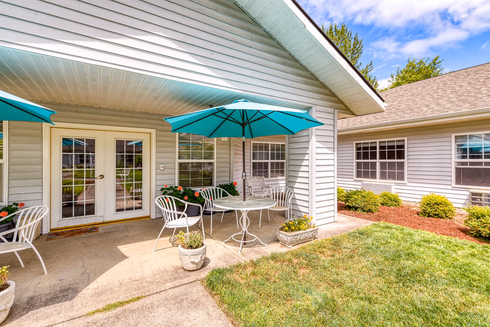 Outdoor patio area at Creekside Place Memory Care with white metal chairs and tables under teal umbrellas, potted plants, and a grassy lawn adjacent to a light gray building with white-framed windows and double glass doors.