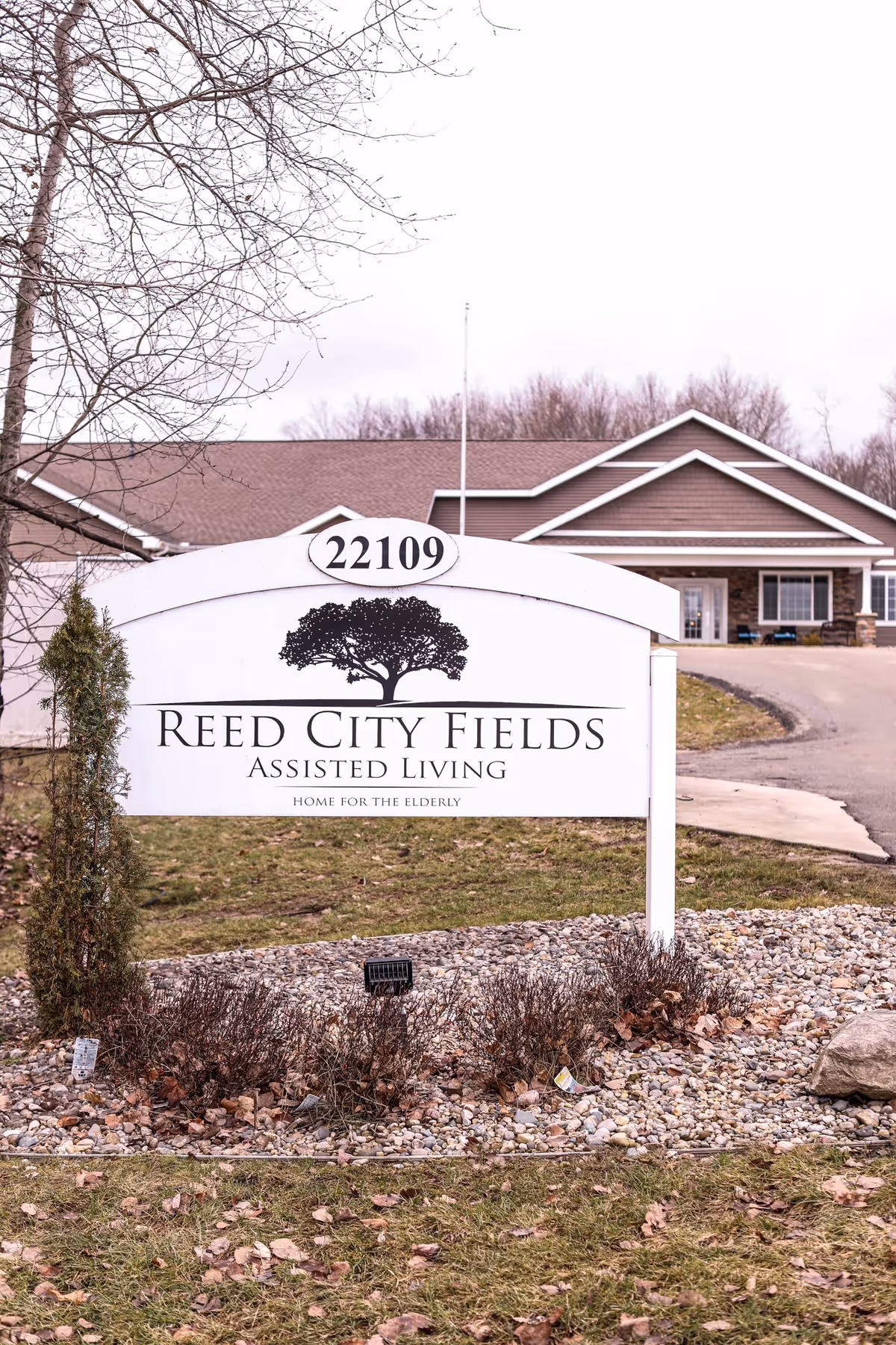 Outdoor view of the entrance sign for Reed City Fields Assisted Living, featuring the address 22109. The sign is white with black text and a tree logo, set in a landscaped area with rocks and shrubs. In the background, there is a building with a brown roof and beige siding, along with a driveway leading up to it.