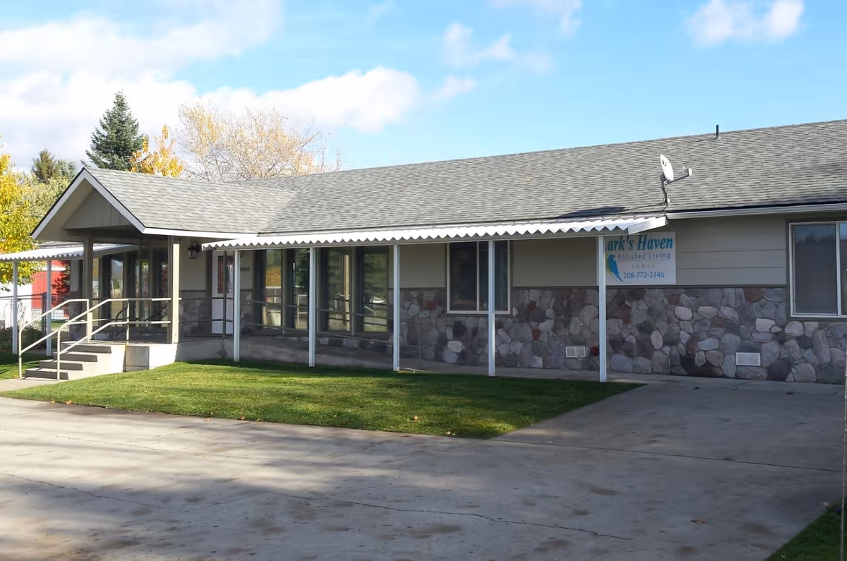 Exterior view of Larkshaven Assisted Living facility showing a single-story building with a stone and siding facade, a covered walkway supported by white posts, a small set of stairs with a handrail leading to the entrance, and a grassy area in front under a partly cloudy sky.