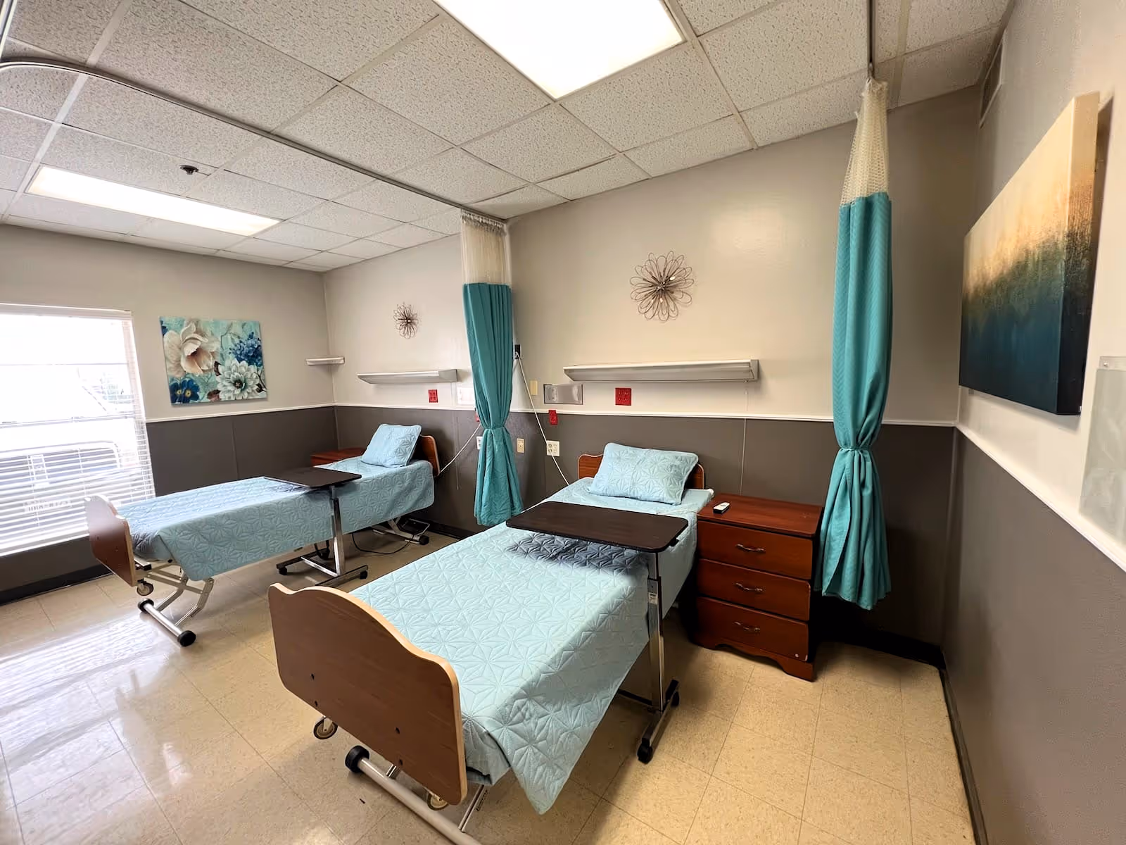 A room with two hospital-style beds separated by teal privacy curtains. Each bed has a light blue patterned bedspread and a small overbed table. There are two wooden nightstands, wall-mounted shelves, and decorative wall art. The room has a large window with blinds and a tiled floor.