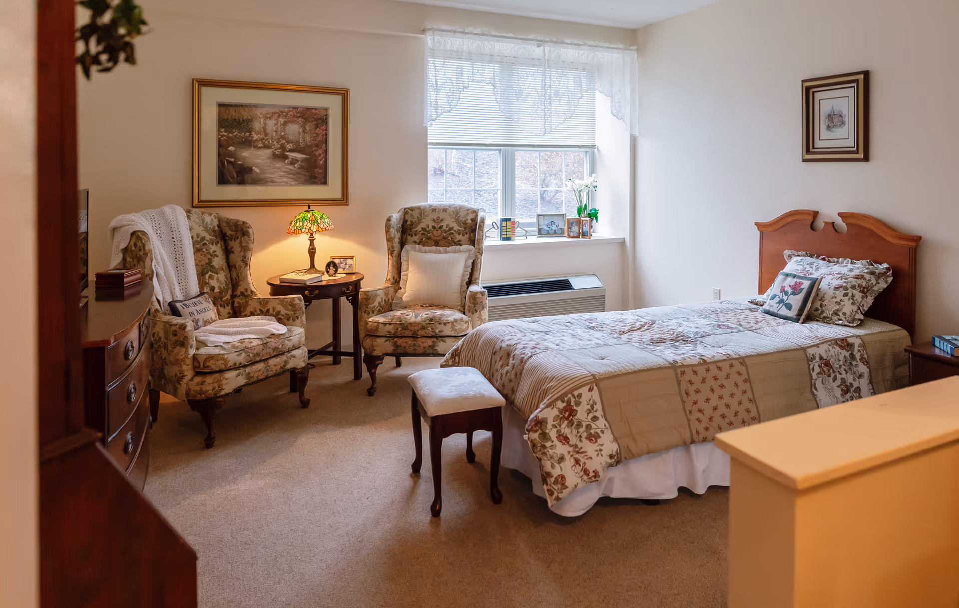 A cozy bedroom with a single bed covered in a floral patchwork quilt. There are two floral upholstered armchairs with a small round wooden table between them, holding a decorative lamp and framed photos. A window with lace curtains lets in natural light, and there are framed pictures on the walls. The room has a warm and inviting atmosphere.