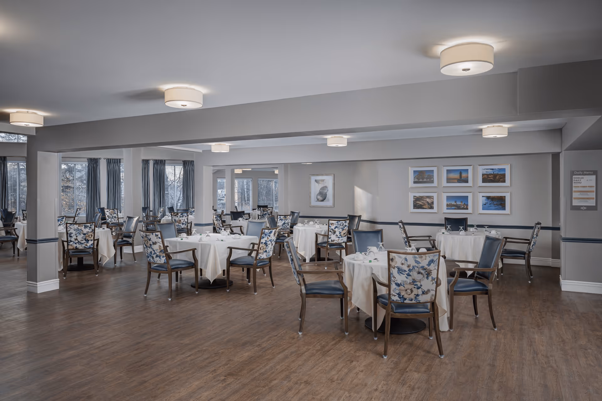 A spacious dining room in a senior living facility with multiple round tables covered with white tablecloths. Each table is surrounded by chairs with blue floral upholstery and dark wooden frames. The room has large windows with blue curtains, light gray walls adorned with framed pictures, and modern ceiling lights. The floor is wooden, and the atmosphere is calm and inviting.