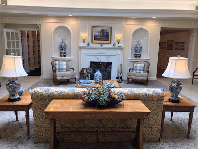 A cozy living room area with a floral patterned sofa facing a white fireplace. On either side of the fireplace are built-in alcoves with large blue and white vases on wooden stands. Two armchairs with plaid cushions are positioned near the fireplace. Two matching table lamps with blue and white bases sit on wooden side tables flanking the sofa. A wooden console table behind the sofa holds a floral arrangement. The room has soft lighting and a framed painting above the fireplace.