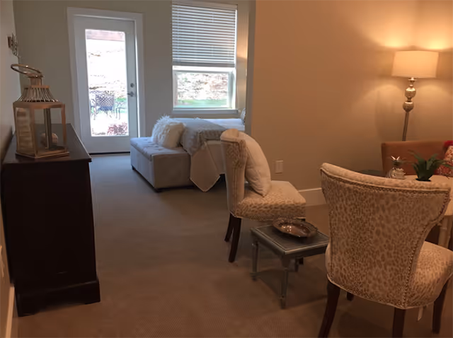 Cozy living room area with upholstered chairs, a small side table and lamp, and a glass door leading outside.