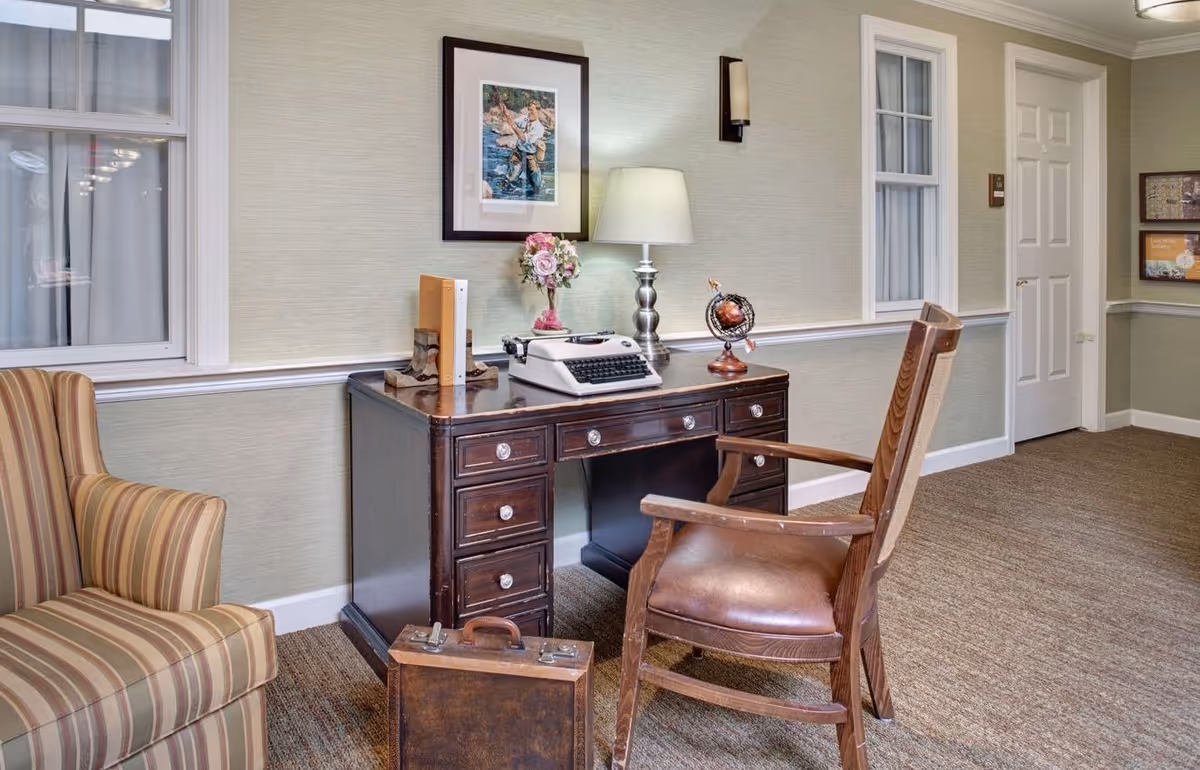 Cozy interior sitting area with a wooden desk holding a typewriter, lamp and globe, a leather desk chair and a striped armchair.