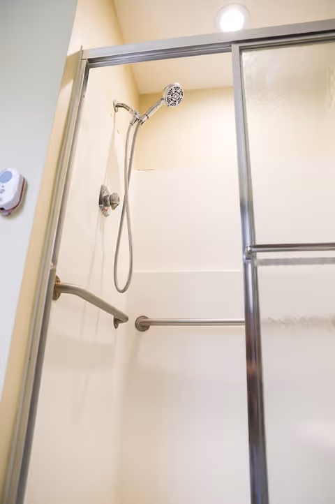 Interior view of a shower stall with a handheld showerhead, grab bars on the walls, and a sliding glass door partially open. The shower area is well-lit with a ceiling light.
