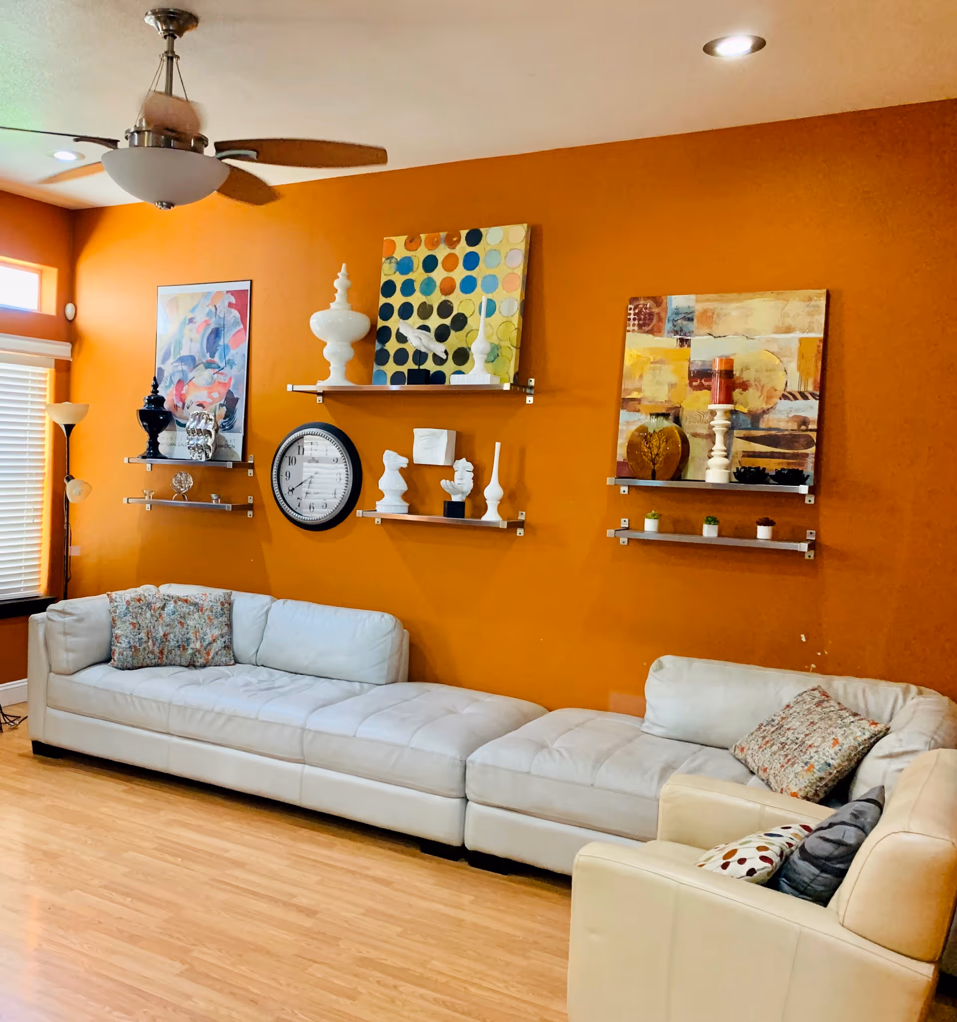 Living room with an orange accent wall, a long white sectional sofa, decorative shelves and colorful artwork.